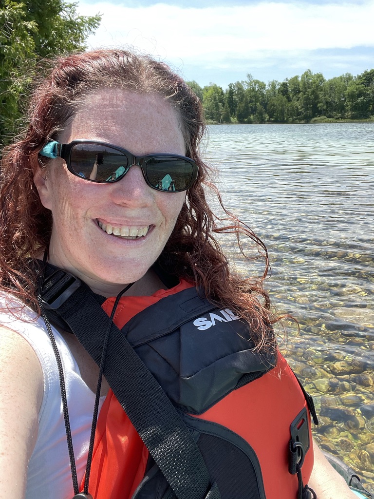 Smiling woman wearing sunglasses and a PFD sitting on the water's edge